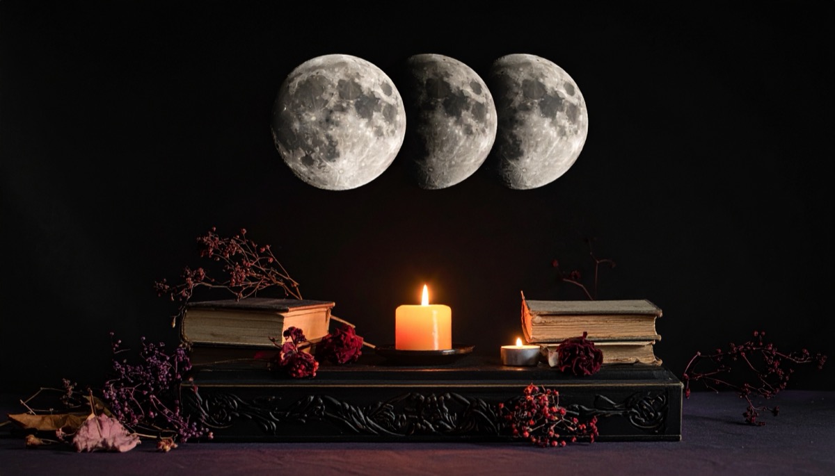three moon phases above a lit candle, old books and dried flowers on a dark altar