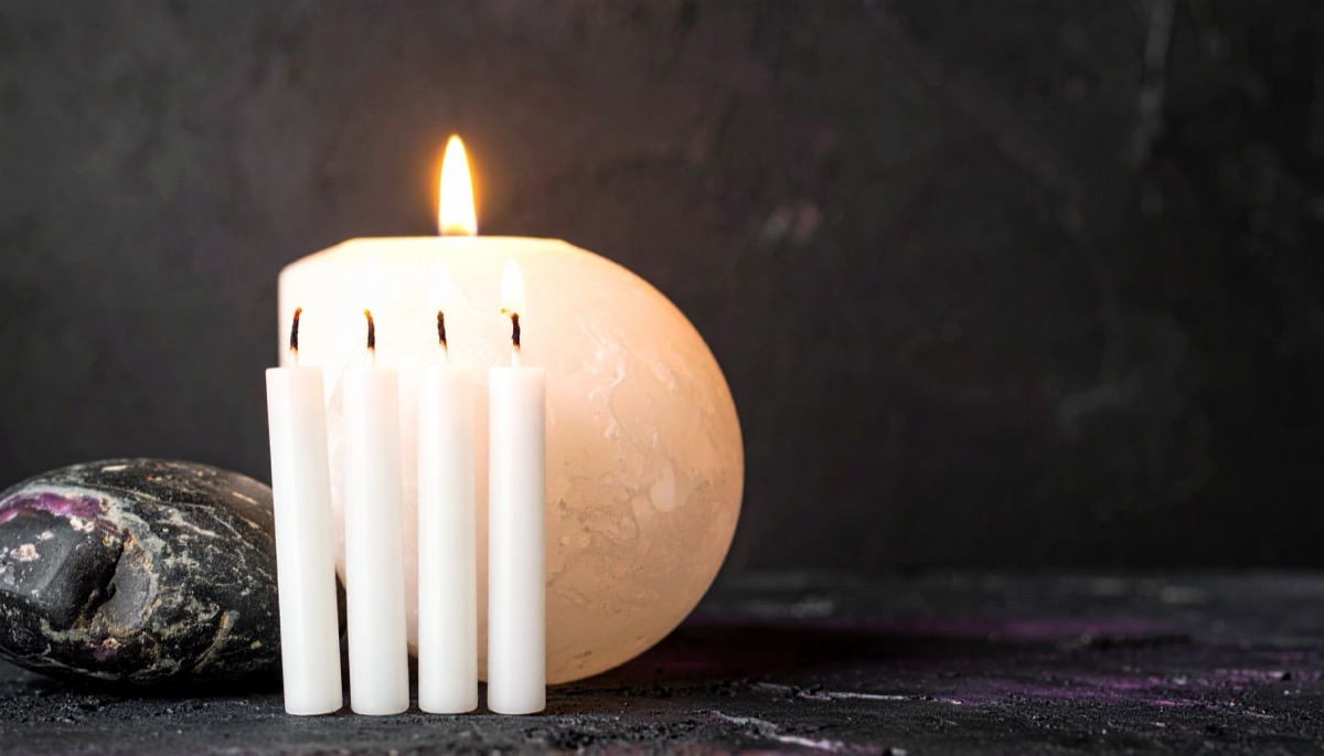 four slim white candles standing before one large lit pillar candle beside a dark stone