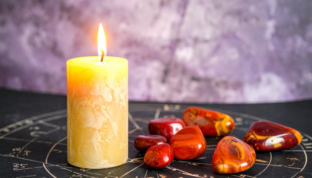 a cluster of red and orange tumbled stones beside a lit cream pillar candle on a zodiac wheel