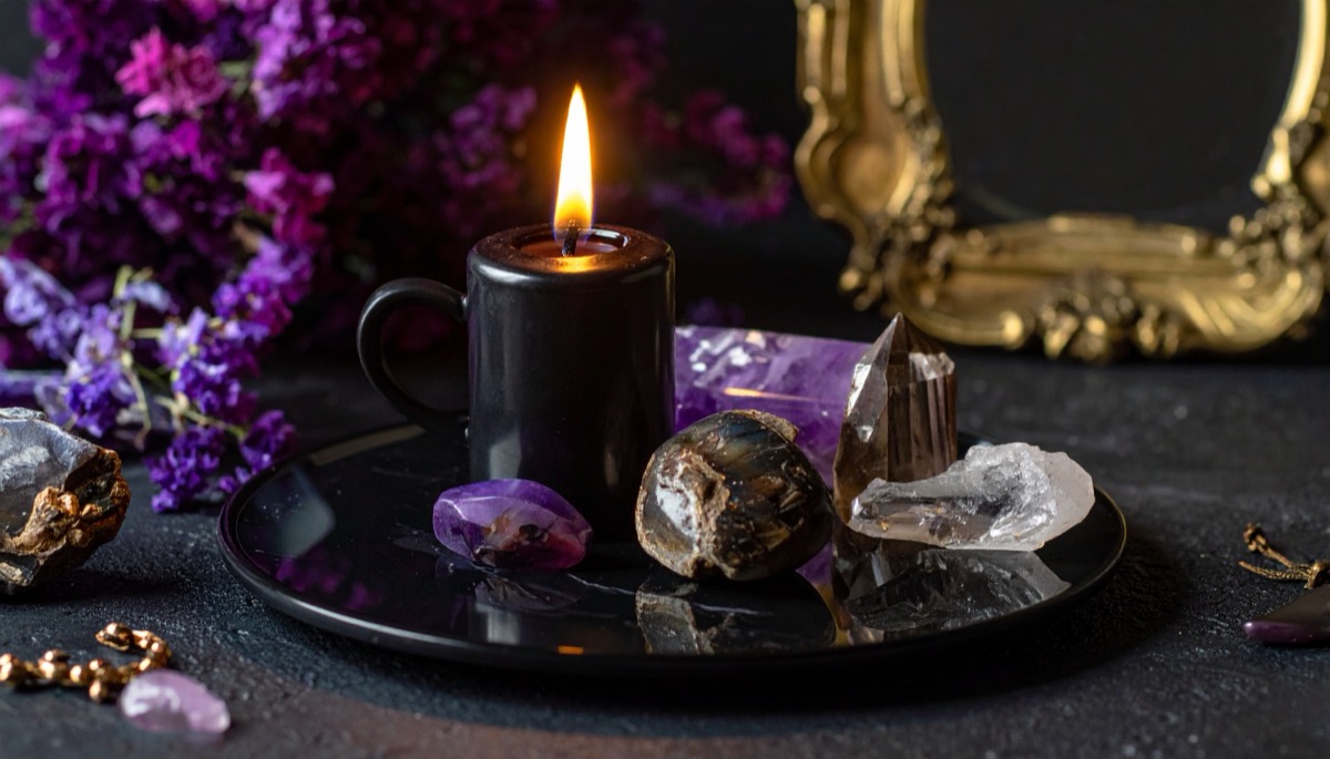 a black candle on a dark tray with violet and clear rough stones beside a gilded frame