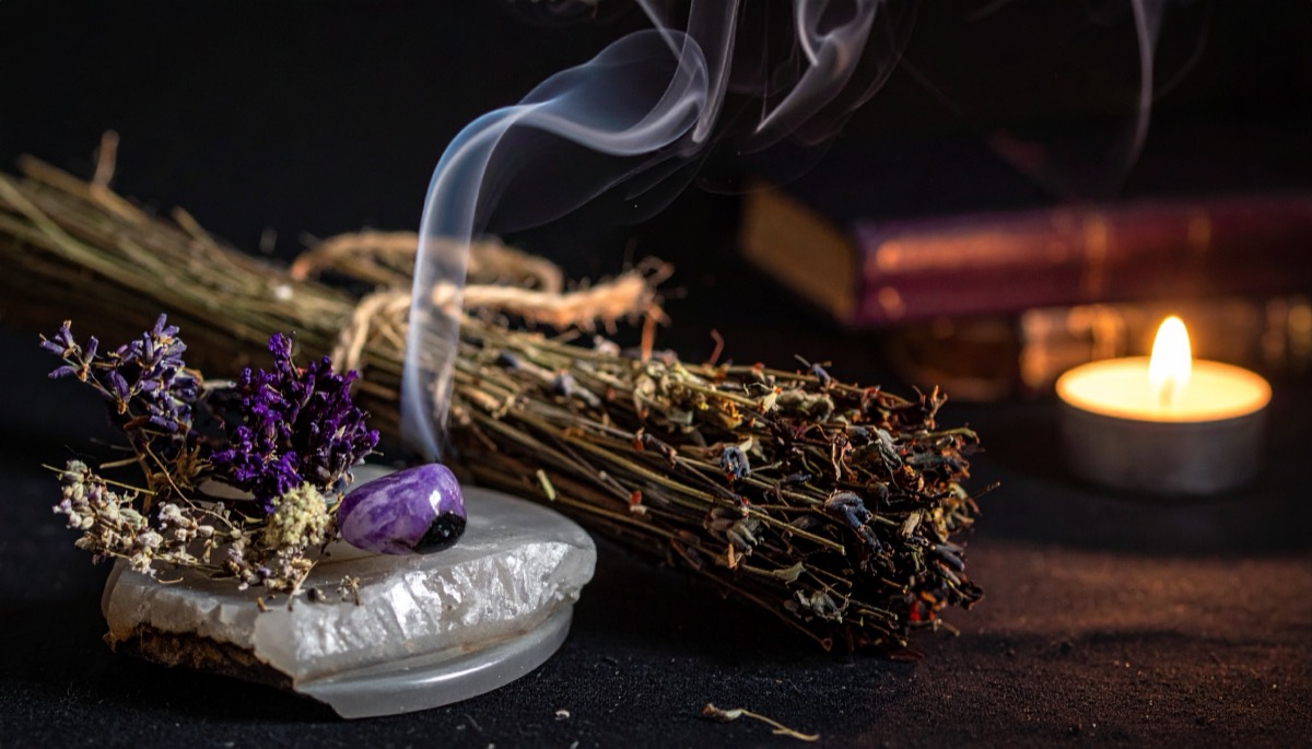 a bundle of dried herbs smoking beside a purple tumbled stone on a white slab